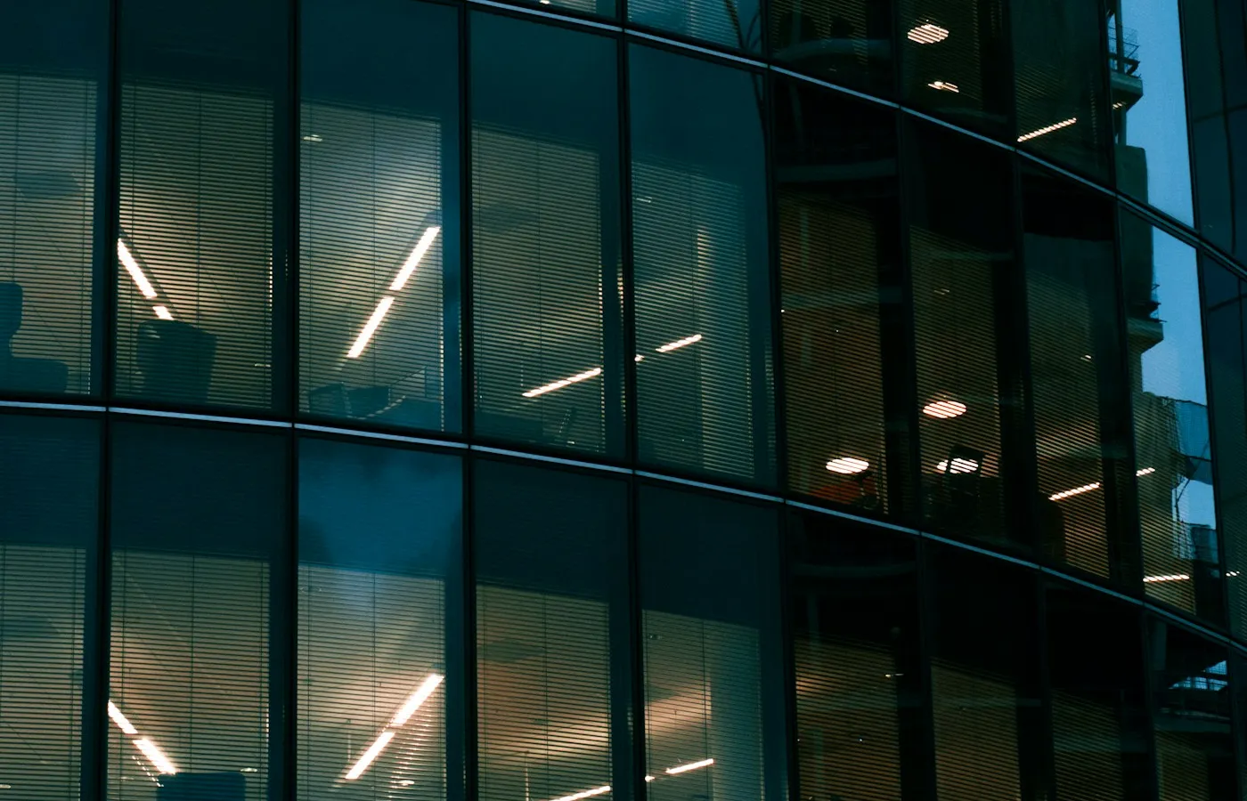 Illuminated office windows with rows of blinds and ceiling lights visible through the glass.